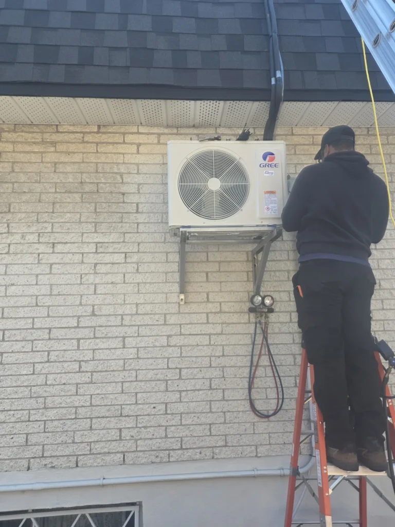 Technicien sur une échelle raccordant l'unité extérieure d'une thermopompe murale Gree sur un mur de brique